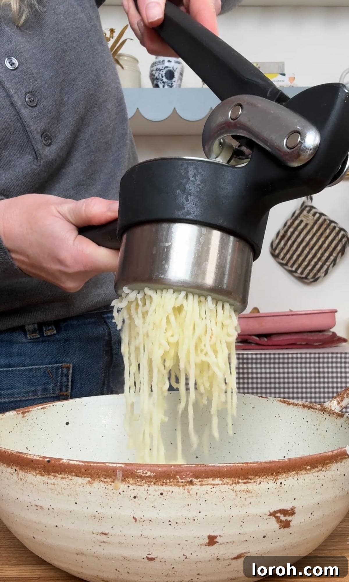 Mashing roasted potato flesh in a bowl with a potato ricer for a smooth texture.