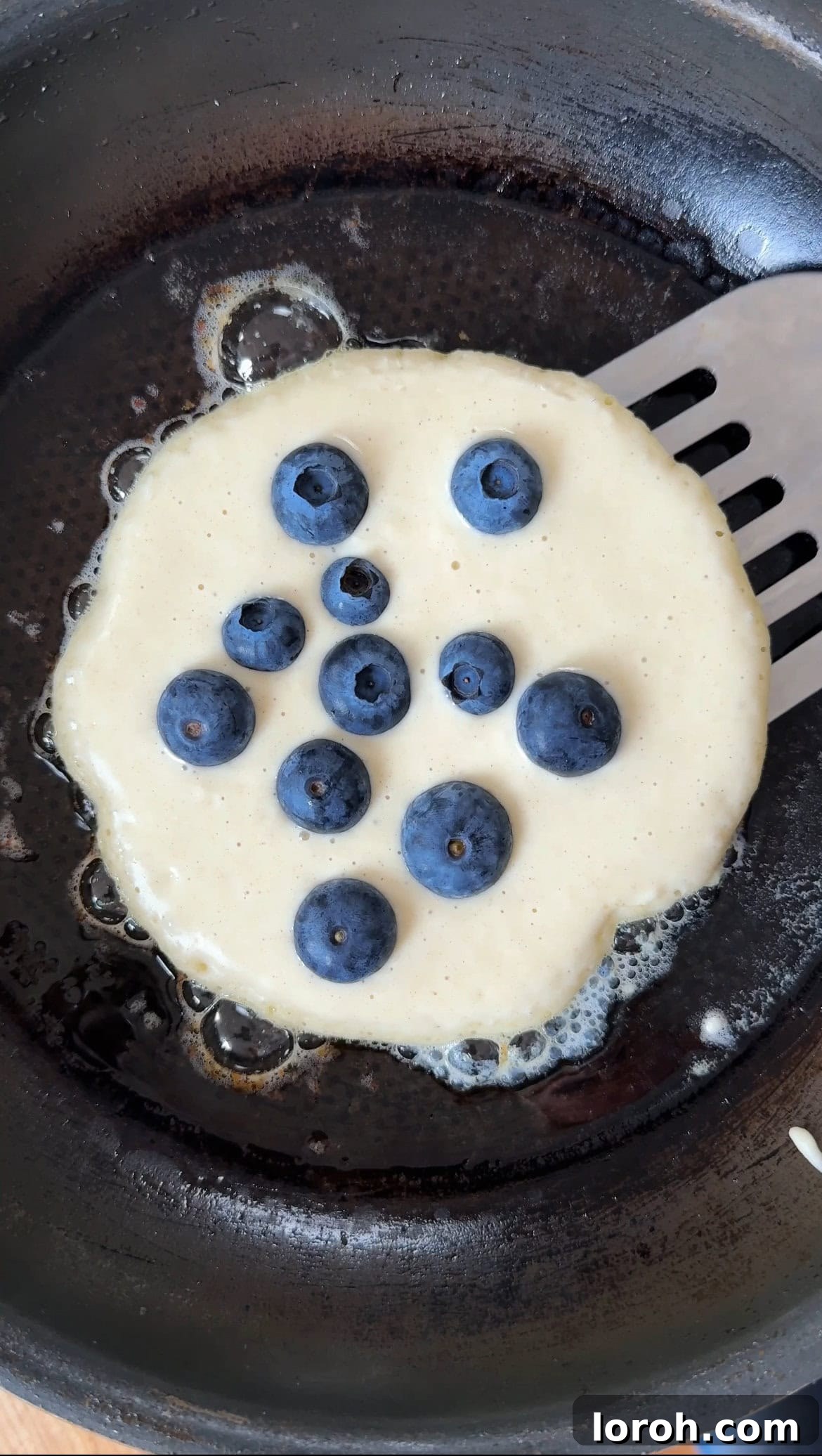 Effortless Fluffy Pancakes Without Baking Powder 6 Blueberries being sprinkled onto pancakes cooking in a pan, before they are flipped.