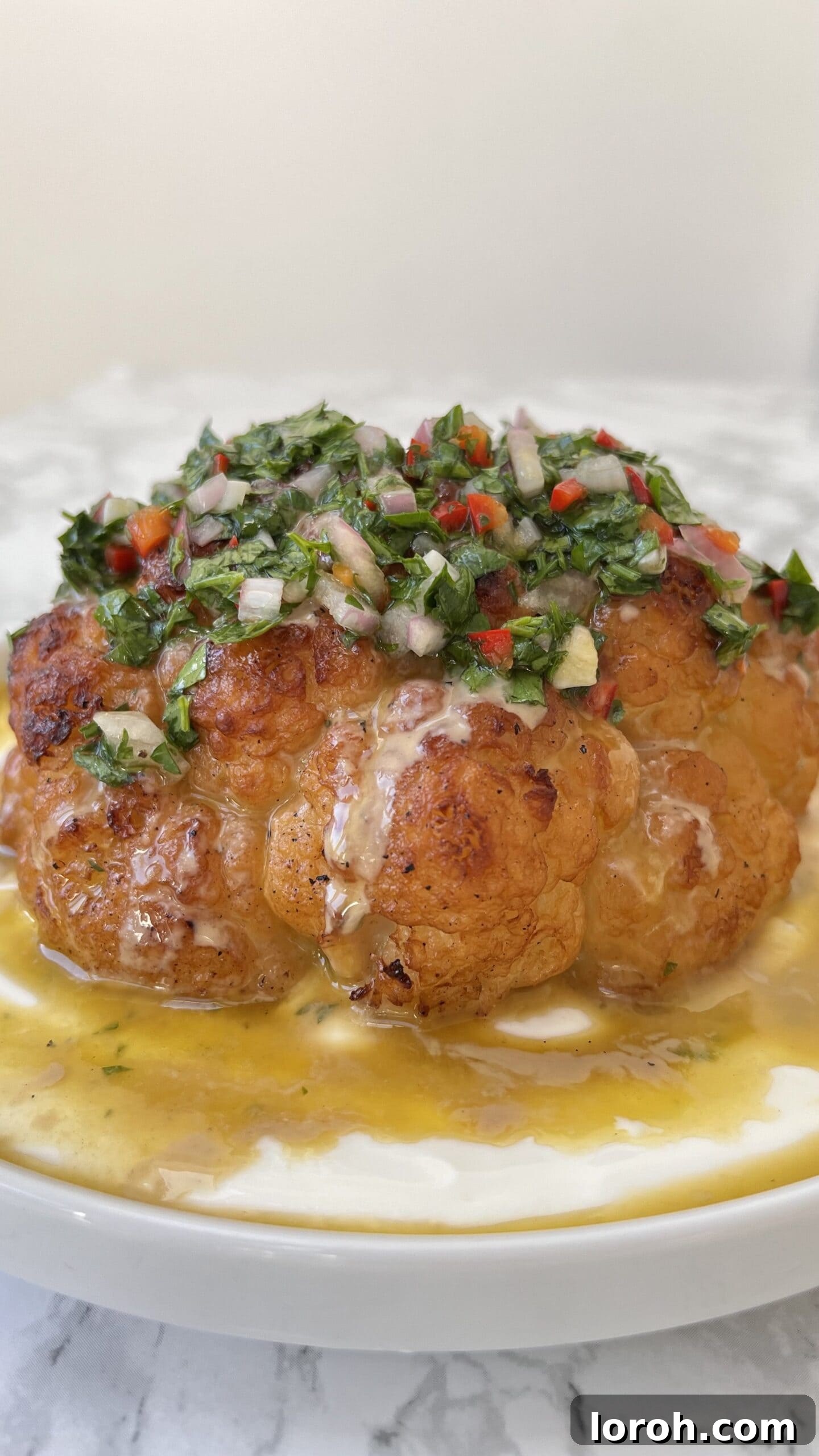 The Ultimate Whole Roasted Cauliflower with Tahini and Chimichurri 2 Close up of a whole roasted cauliflower with tahini sauce and chimichurri spooned over the top, showcasing its golden char and vibrant toppings.