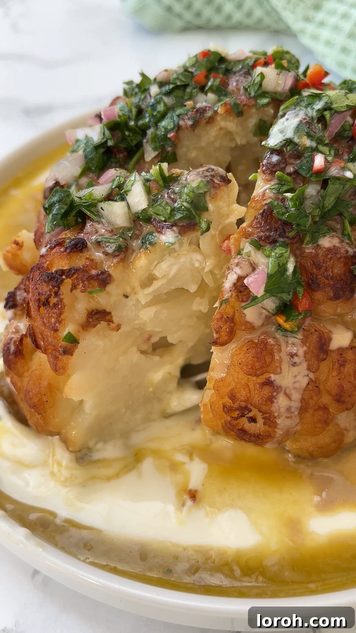 The Ultimate Whole Roasted Cauliflower with Tahini and Chimichurri 3 A beautifully roasted cauliflower with tahini and chimichurri, with a wedge already removed, highlighting its tender interior and crispy exterior.