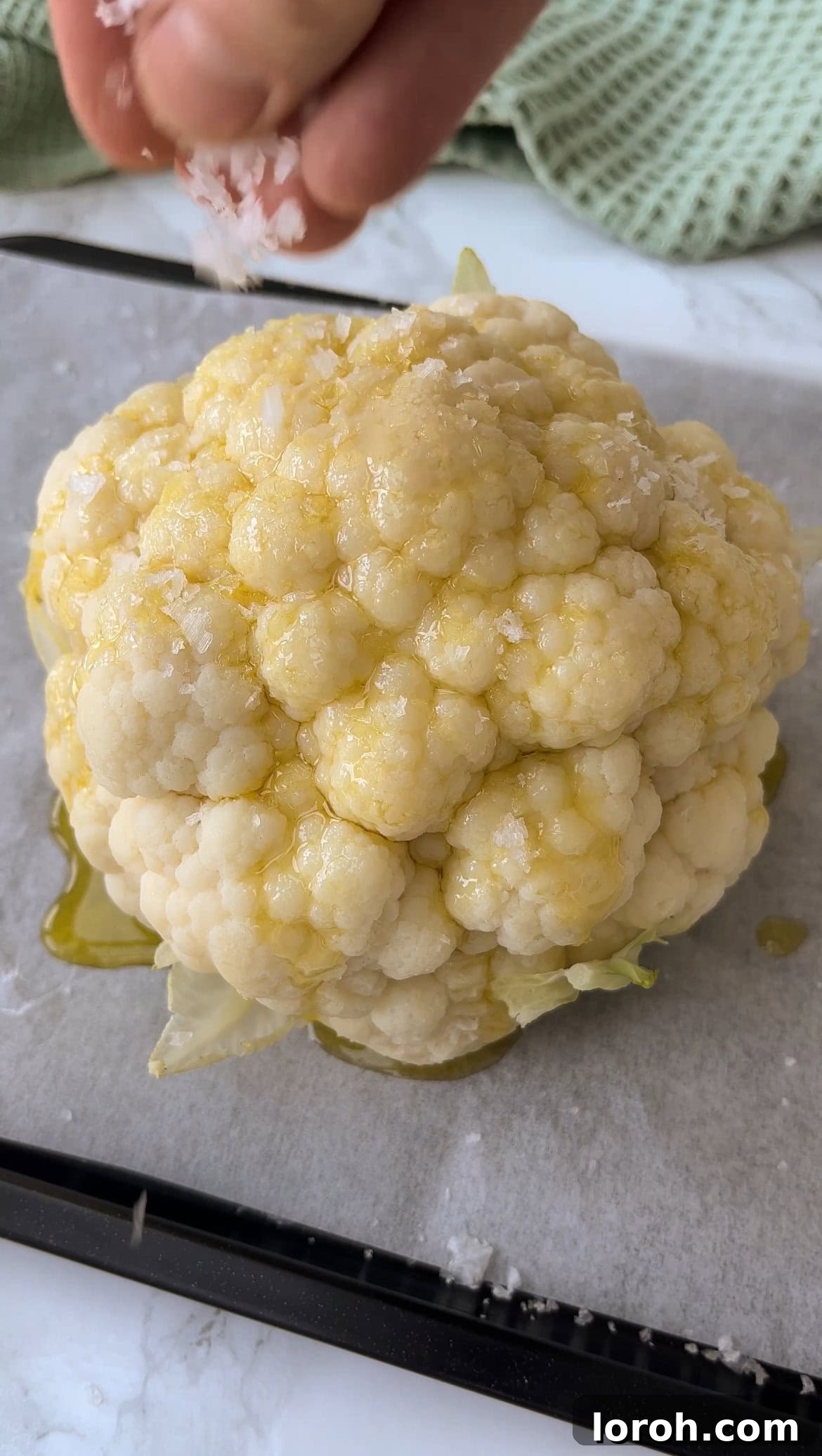 The Ultimate Whole Roasted Cauliflower with Tahini and Chimichurri 5 A parboiled cauliflower on a baking tray, being drizzled with olive oil and seasoned with sea salt before roasting.
