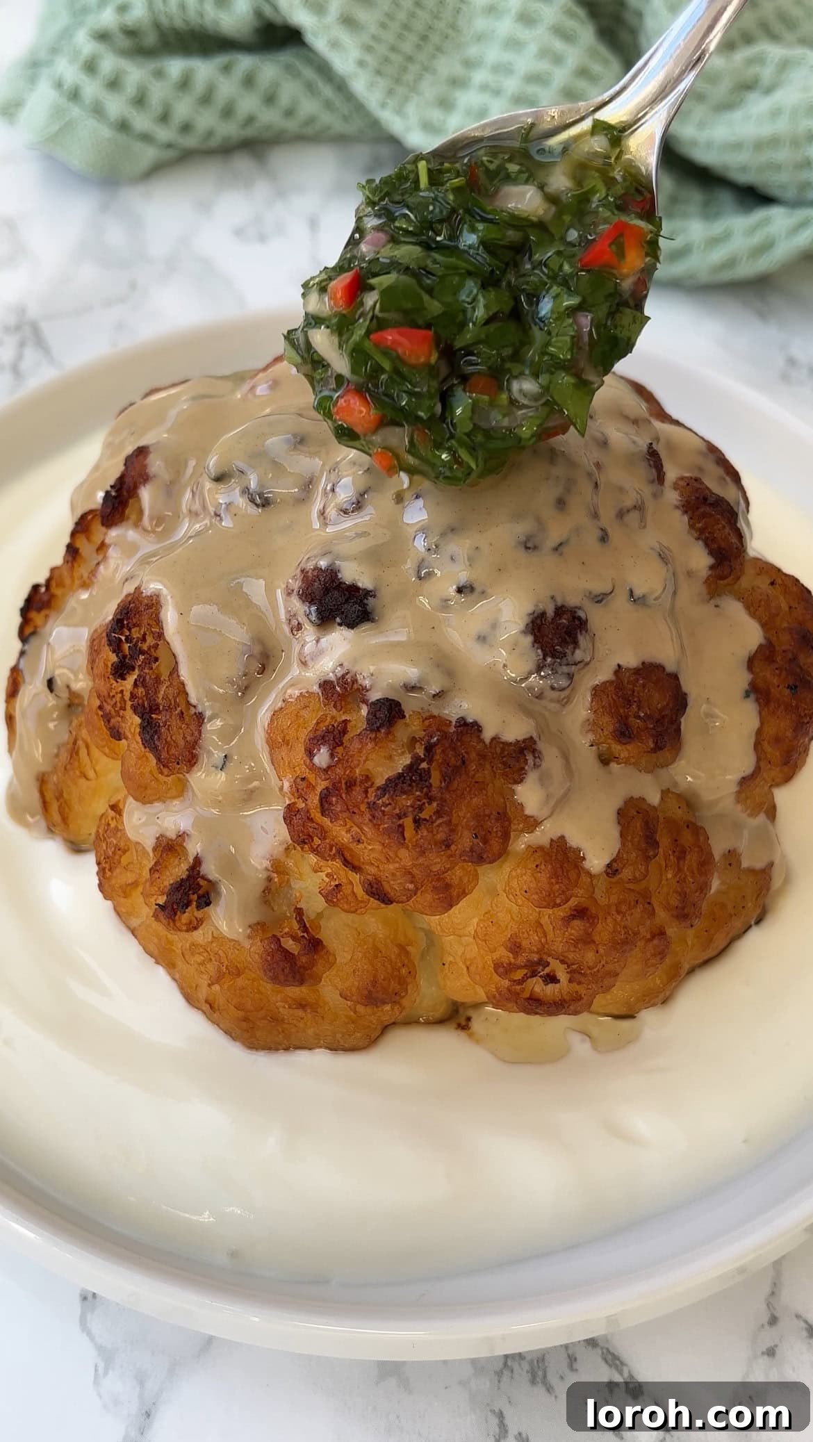 The Ultimate Whole Roasted Cauliflower with Tahini and Chimichurri 8 Close up of a whole roasted cauliflower, with vibrant chimichurri being added over the tahini sauce, creating a beautiful contrast.