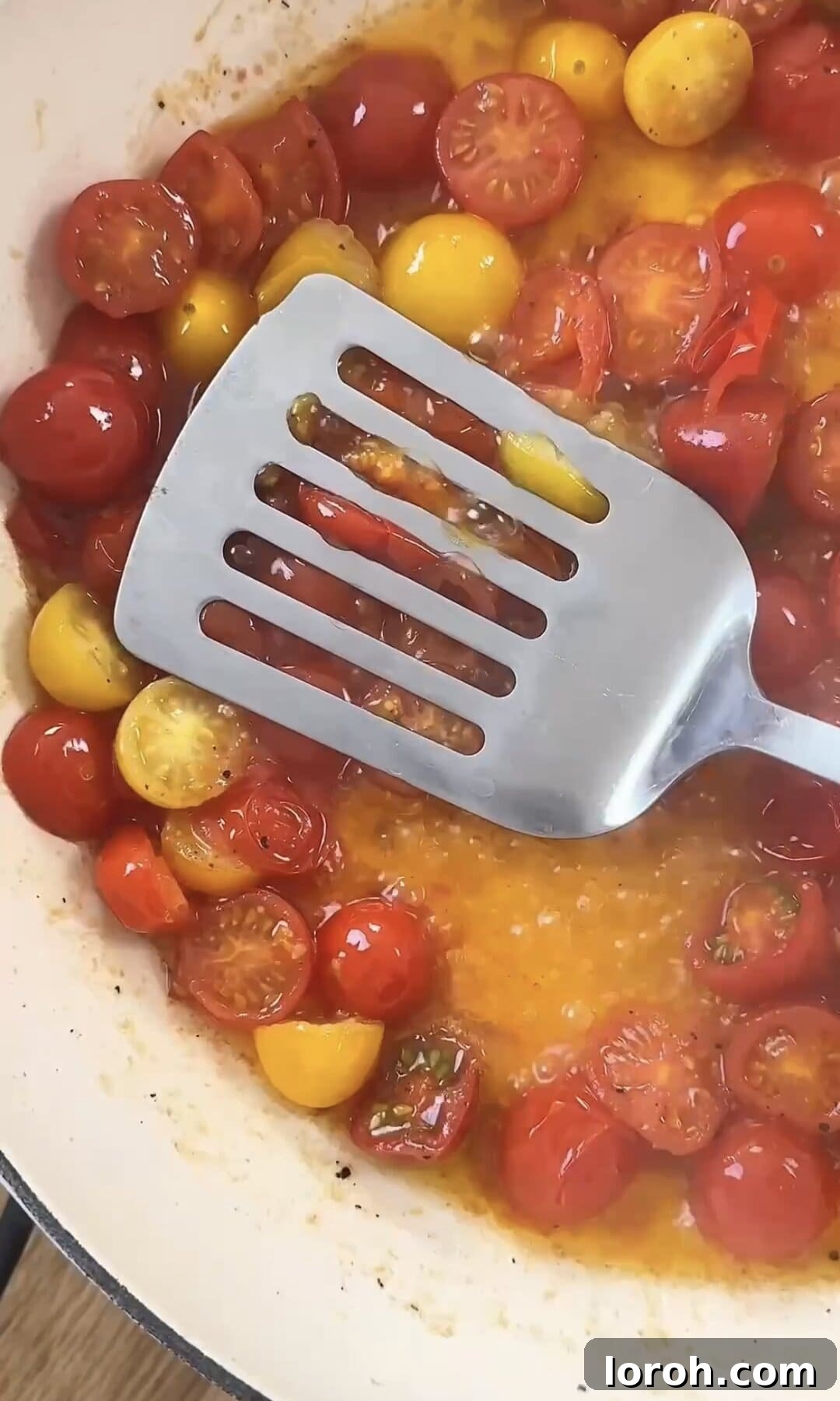 Using a spatula to gently press down on bursting cherry tomatoes in a pan, forming a sauce.