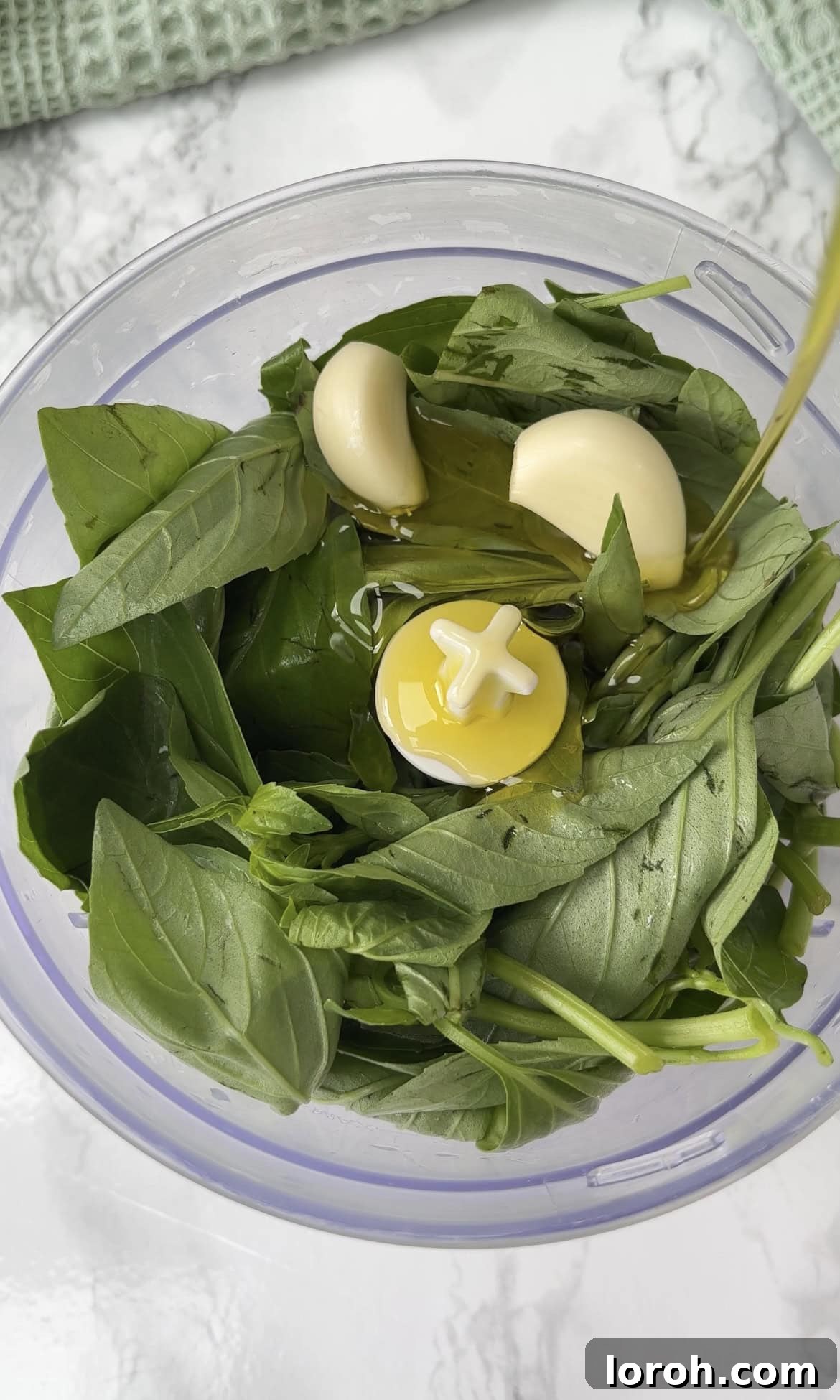 Fresh basil leaves being added to a food processor with toasted pine nuts, garlic, and olive oil for homemade pesto.