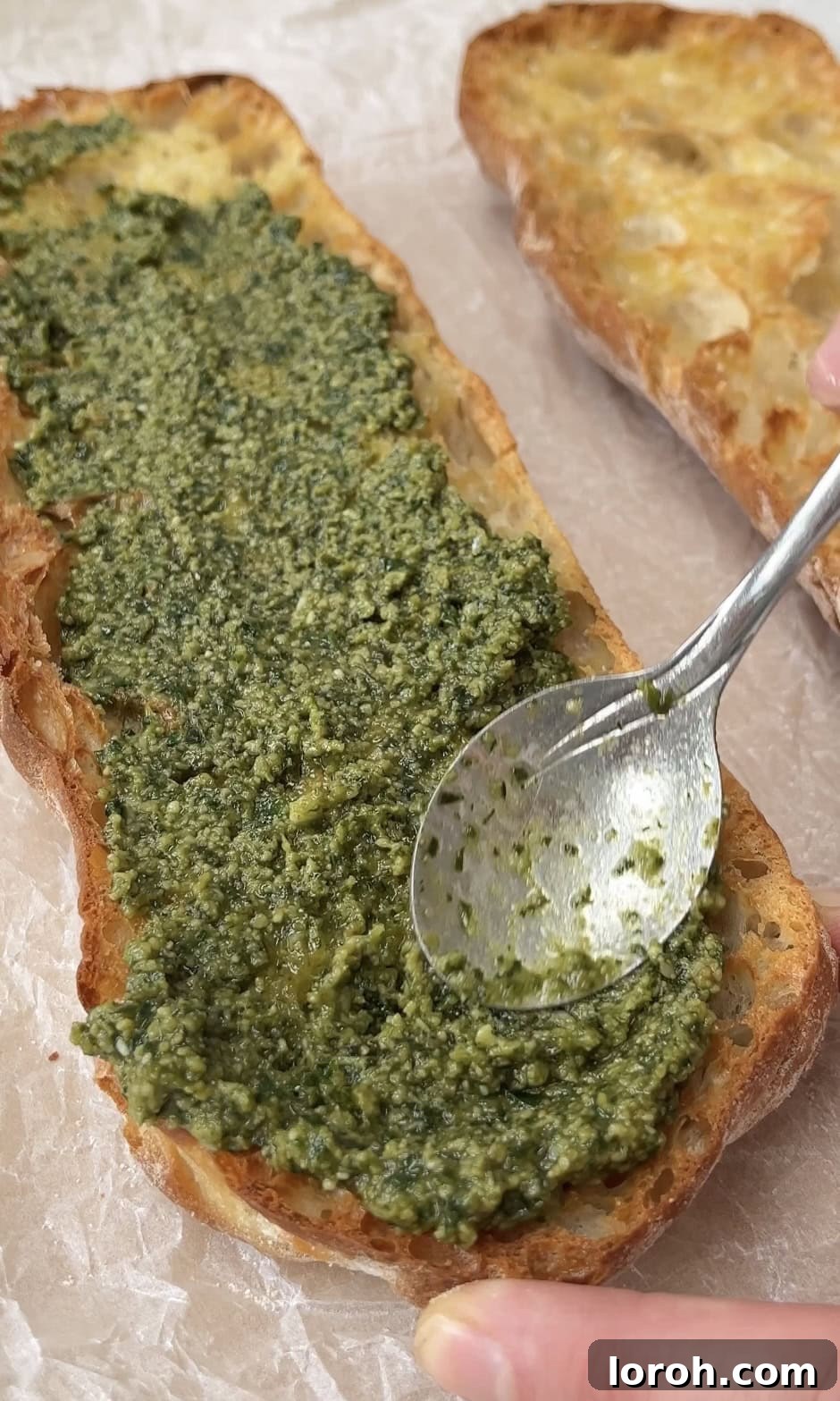 Freshly made basil pesto being generously spread onto a toasted, garlic-rubbed ciabatta slice.