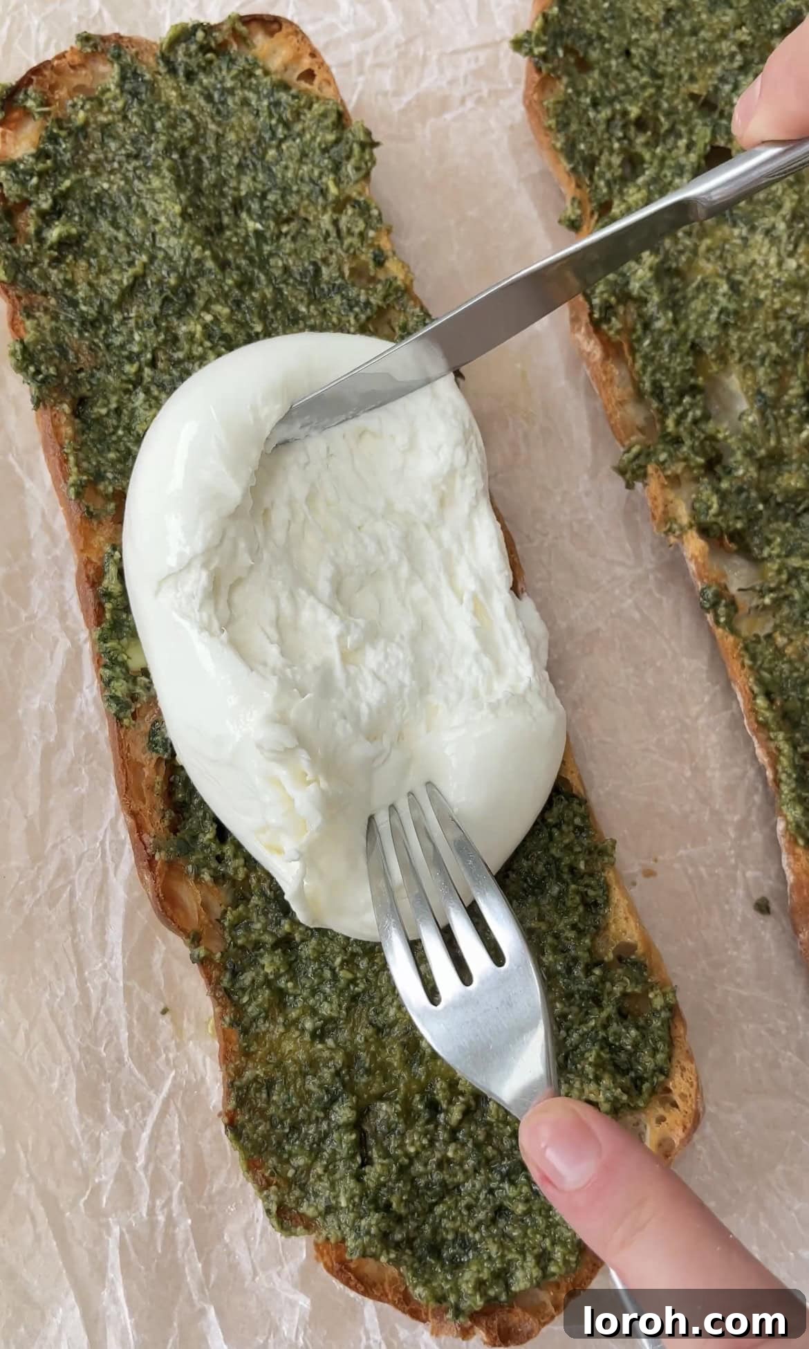 Creamy burrata cheese being gently smoothed and spread over a pesto-coated ciabatta slice.