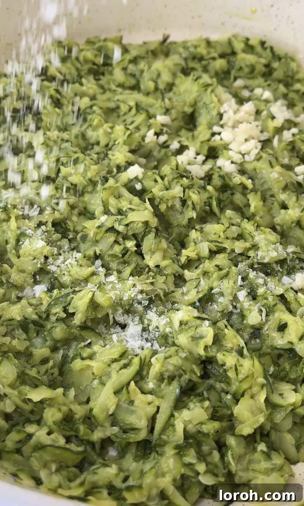 Crushed garlic cloves being added to the softened courgettes in a pan, releasing their aroma.
