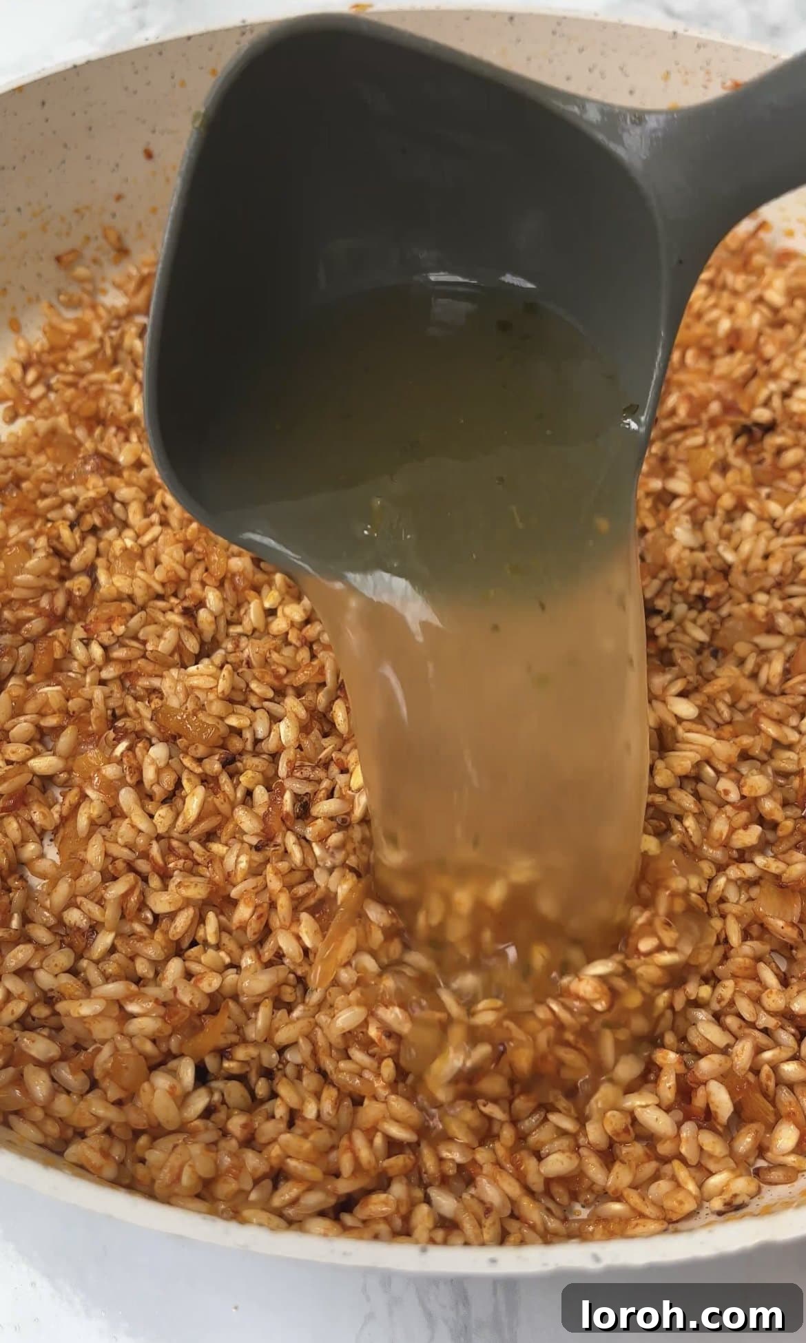 Adding a ladleful of hot vegetable stock to the toasted risotto rice, stirring gently.
