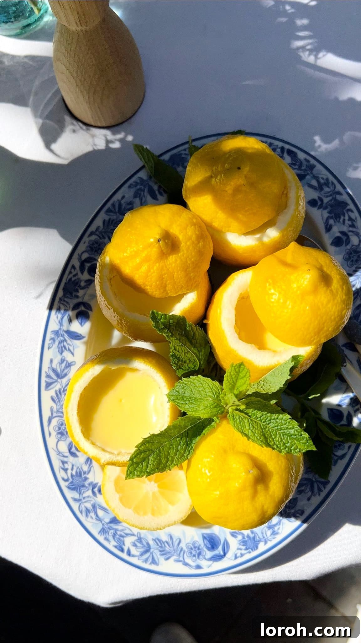 Luscious Lemon Cream 2 Two lemon possets served in scooped-out lemon halves, garnished with mint leaves, resting on a blue plate.