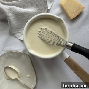 A whisk in a saucepan of creamy white sauce, showing its smooth and glossy texture.