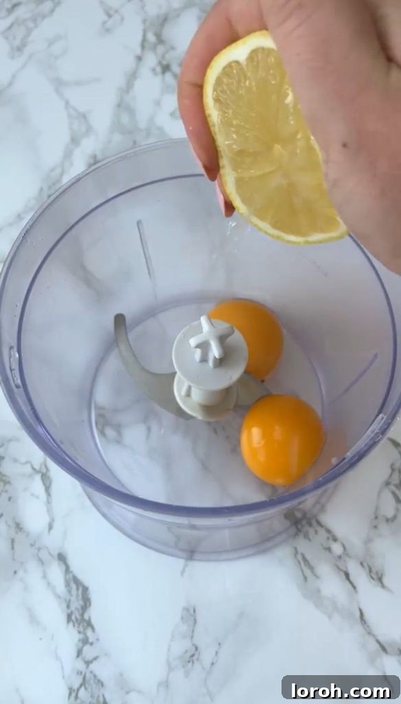 Two fresh egg yolks in a blender, with a hand adding lemon juice from half a lemon