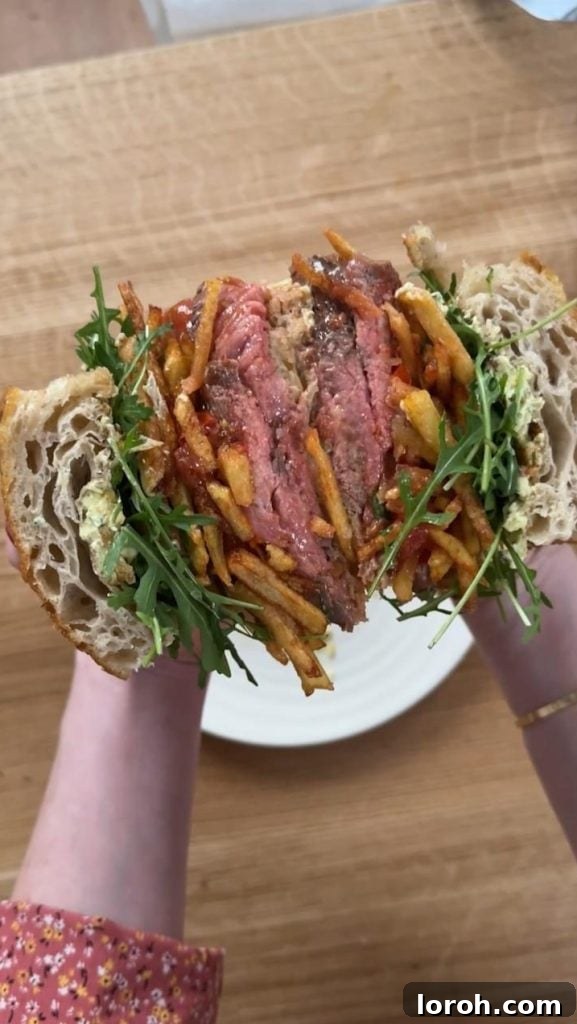 A woman proudly holding a stunning rib eye steak sandwich, expertly layered with succulent steak slices, crispy homemade chips, glistening jammy tomatoes, fresh rocket, and a dollop of creamy bearnaise butter. The gourmet sandwich is presented on a wooden board, inviting viewers to savor its rich textures and flavors.