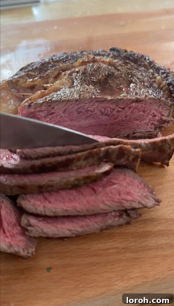 A chef's knife expertly slicing a perfectly rested medium-rare rib eye steak, revealing a vibrant pink center and juicy texture. The cut is clean, indicating a tender piece of meat, with juices glistening on the cutting board.