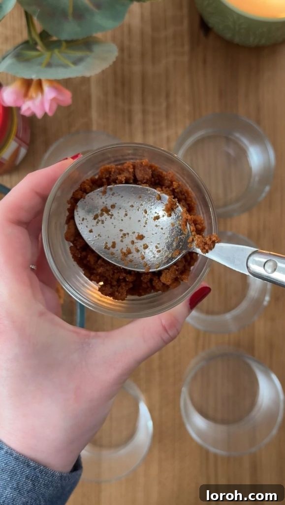 Five Ingredient No Bake Biscoff Cheesecake 5 A woman meticulously packing the cheesecake base into a glass using a spoon, demonstrating proper technique.