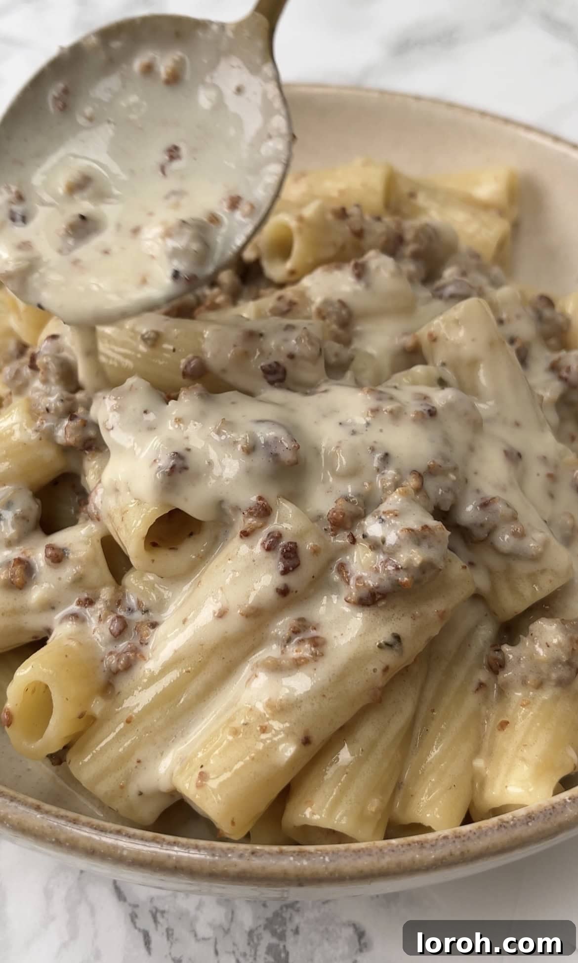 Spoonful of rich, creamy sausage pasta being served, showcasing the perfectly coated rigatoni and savory sausage crumbles, garnished with black pepper.