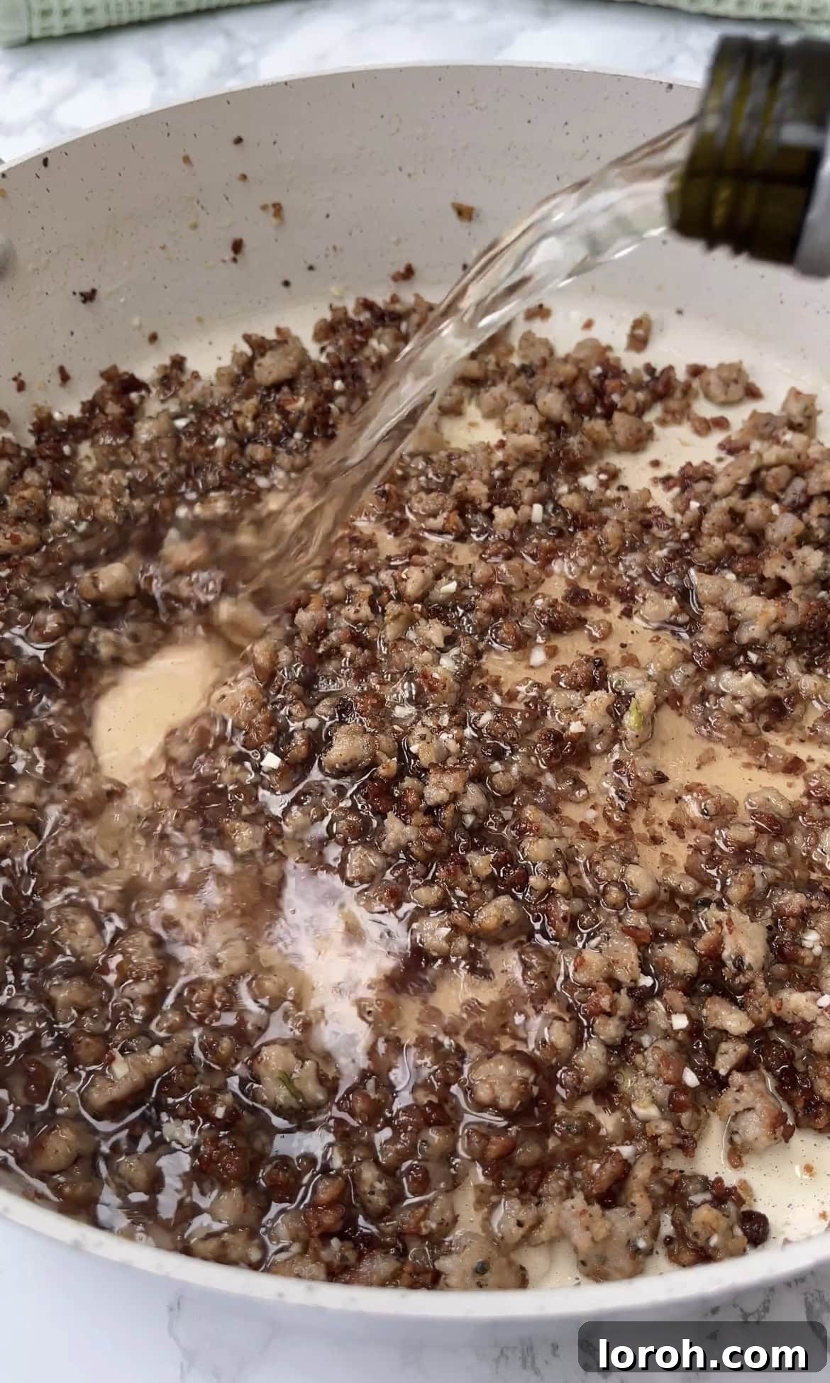 White wine being poured into a pan with browned sausage and fragrant garlic, creating steam as it deglazes the pan.