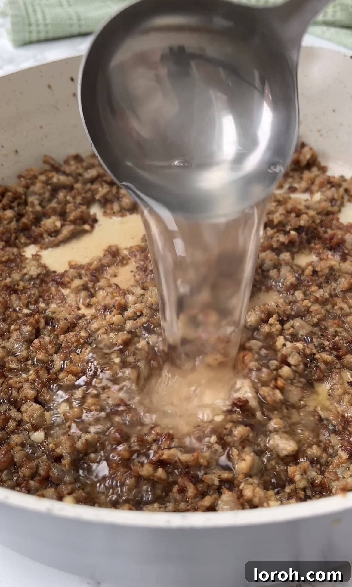 A ladle of starchy pasta water being carefully added to the simmering creamy sausage sauce in a pan.