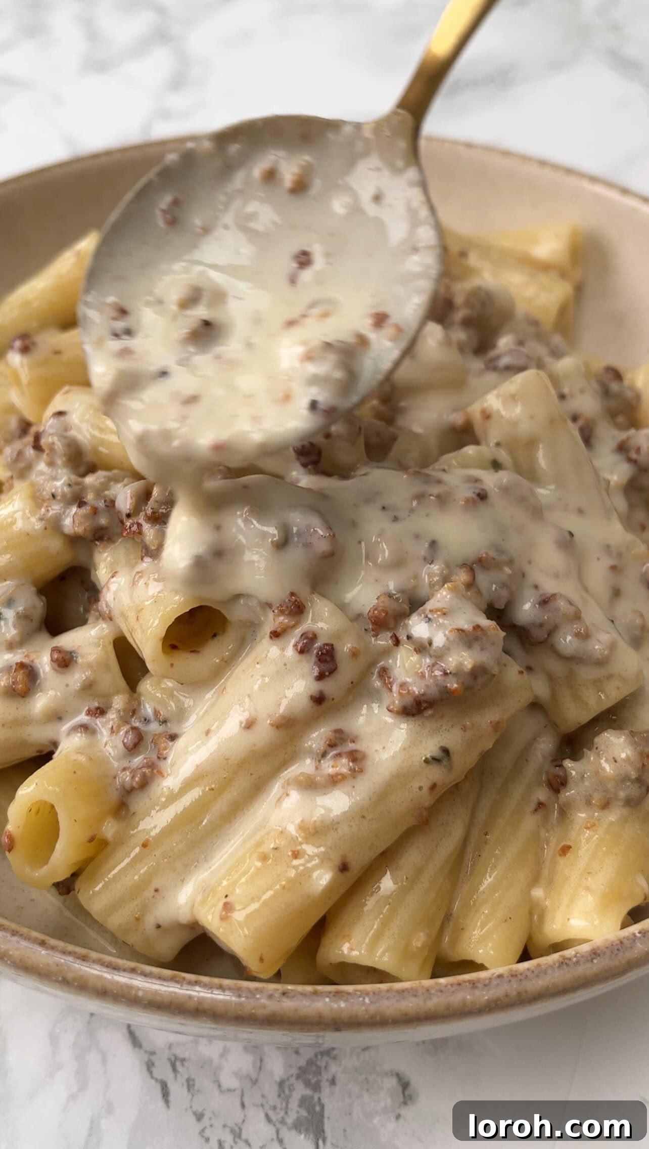 Close-up of a generous serving of creamy sausage pasta in a rustic beige bowl, with sauce being spooned over and a hint of fresh herbs, evoking comfort.
