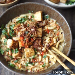 Bowl of curry udon topped with tofu and crispy shallots being eaten with chopsticks.