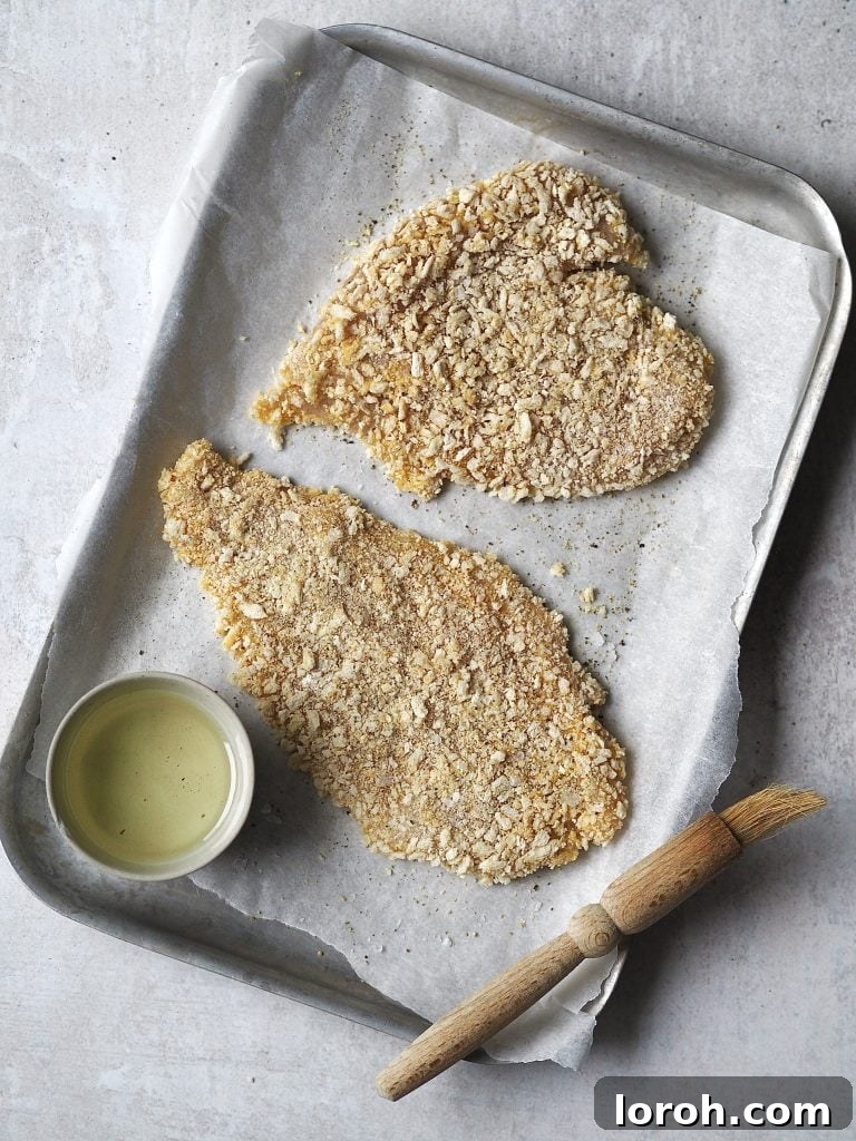 Crispy Chicken with Mediterranean White Bean Salad 7 Breaded chicken cutlets on a lined baking tray being brushed with a bowl of oil.