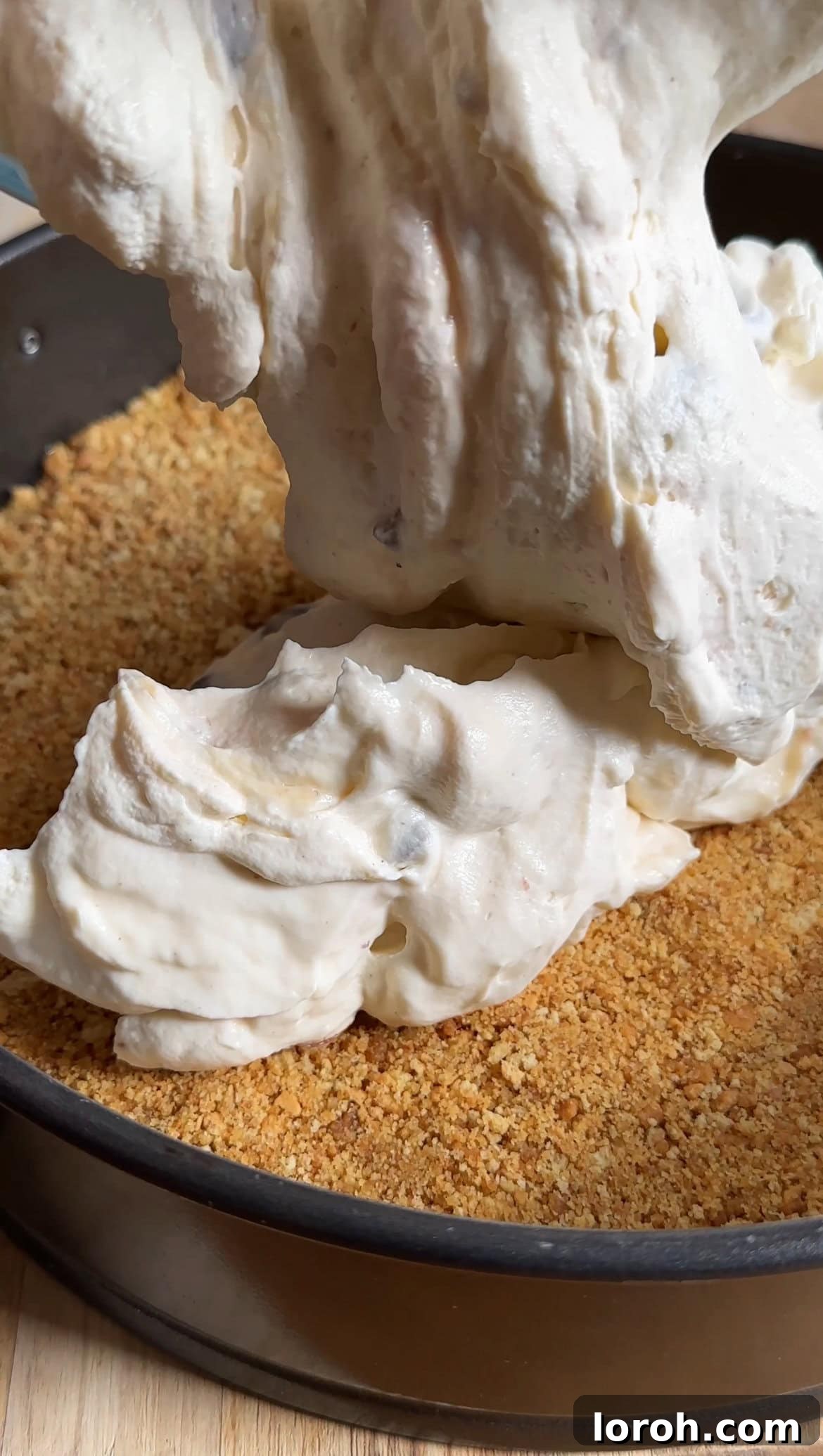 Spoonfuls of creamy Mini Egg cheesecake filling being carefully placed onto the chilled biscuit base in a springform tin, ready to be smoothed.