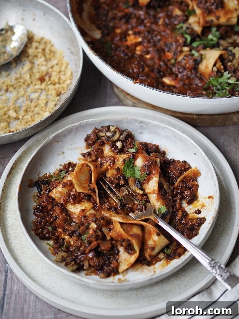 Bowl of hearty vegan lentil bolognese with wide pappardelle pasta, garnished with fresh herbs and a sprinkle of pangrattato.