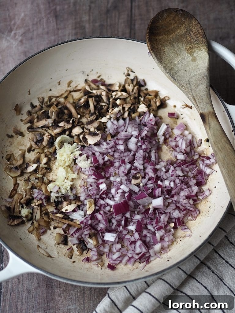 Finely chopped red onion, mushrooms, and minced garlic being gently fried in olive oil in a shallow casserole dish until softened and fragrant, forming the aromatic base of the bolognese.