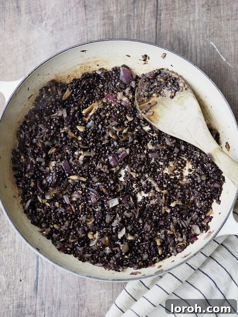 Pre-cooked beluga lentils being stirred into the sautéed onion and mushroom mixture in a shallow casserole dish, absorbing the aromatic flavors and deepening the sauce's foundation.