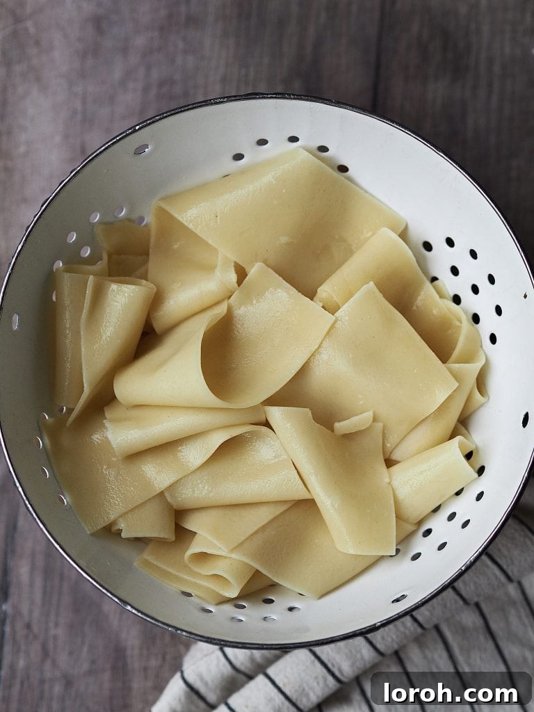 Cooked lasagne sheets, perfectly al dente, resting in a white metal sieve after being drained, ready to be sliced into elegant pappardelle ribbons.