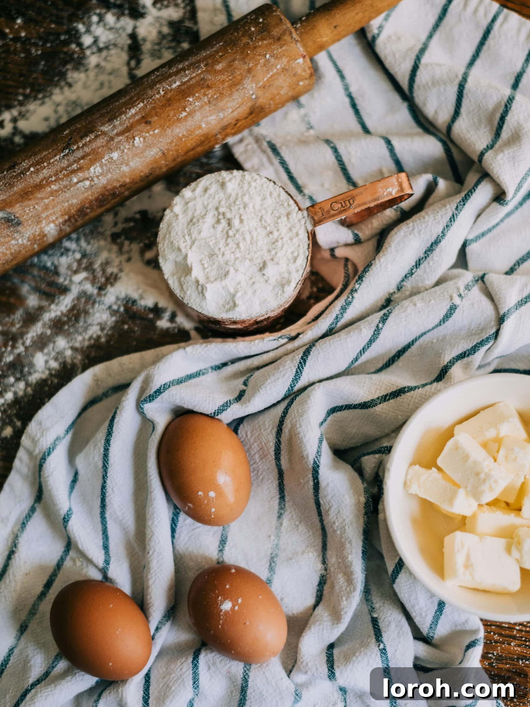 Grams and Ounces Conversion Simplified 2 A beautifully arranged photo of fresh eggs, a measuring cup filled with flour, and softened butter on a rustic tea towel, symbolizing the essentials of precise baking measurements.