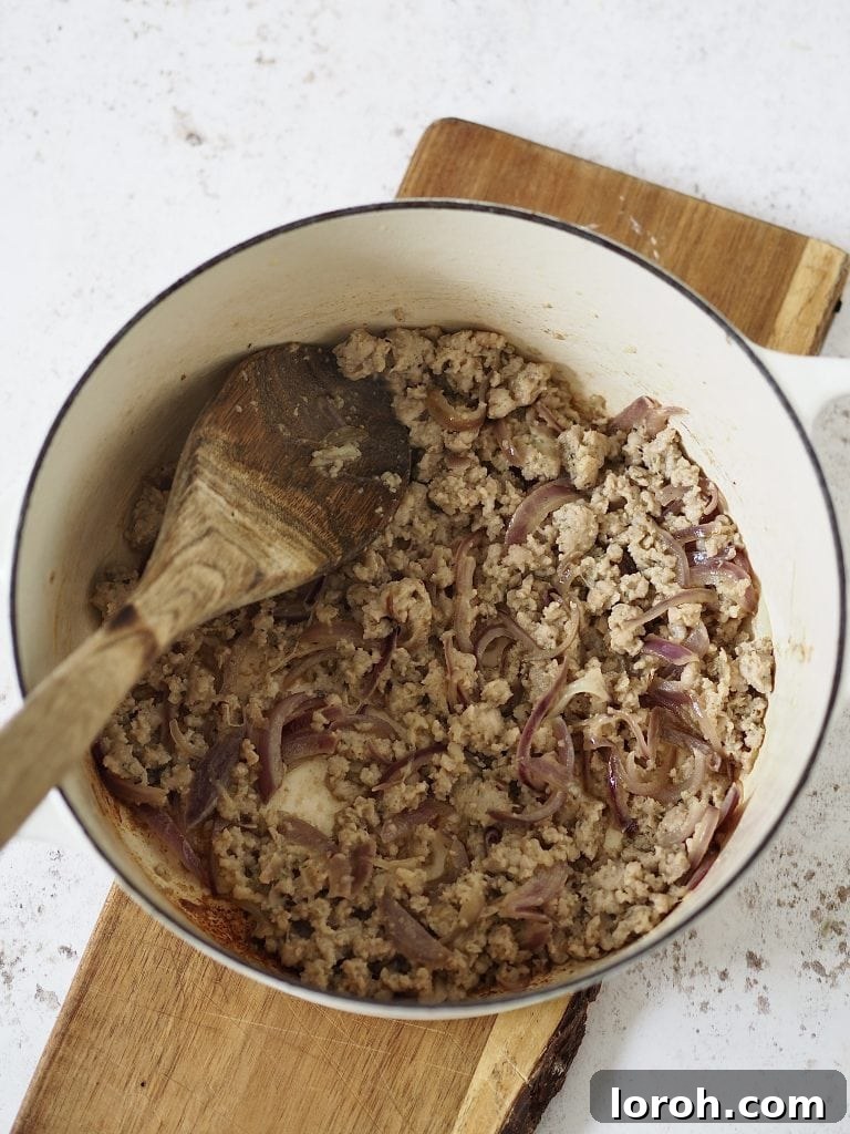 Hearty Weeknight Sausage Ragu 5 Crumbled sausage meat and finely diced red onion sizzling in olive oil within a white casserole dish, starting the flavorful base of the ragu.