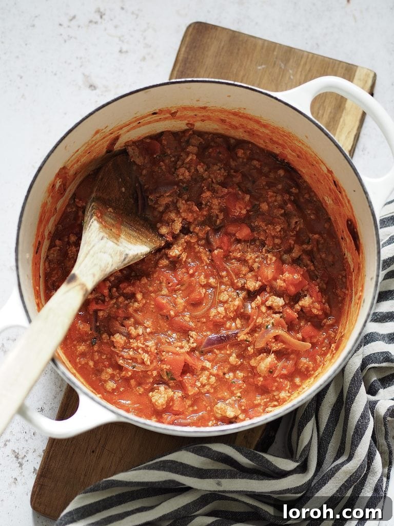 Hearty Weeknight Sausage Ragu 6 The rich sausage ragu bubbling gently in a white casserole dish, stirred with a wooden spoon, indicating it's nearly ready to be combined with pasta.
