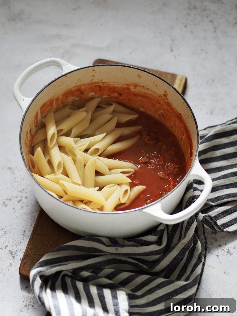 Hearty Weeknight Sausage Ragu 7 Perfectly cooked penne pasta rests atop a bubbling pot of tomato and sausage sauce, waiting to be tossed together for a complete dish.
