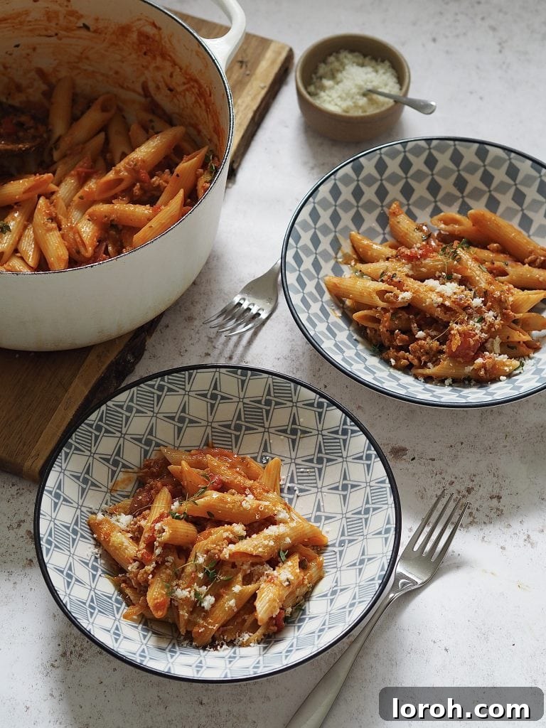 Hearty Weeknight Sausage Ragu 8 Two blue and white bowls of hearty penne pasta with tomato sausage ragu, generously topped with grated cheese, next to a large white casserole dish of more pasta, ready for serving.