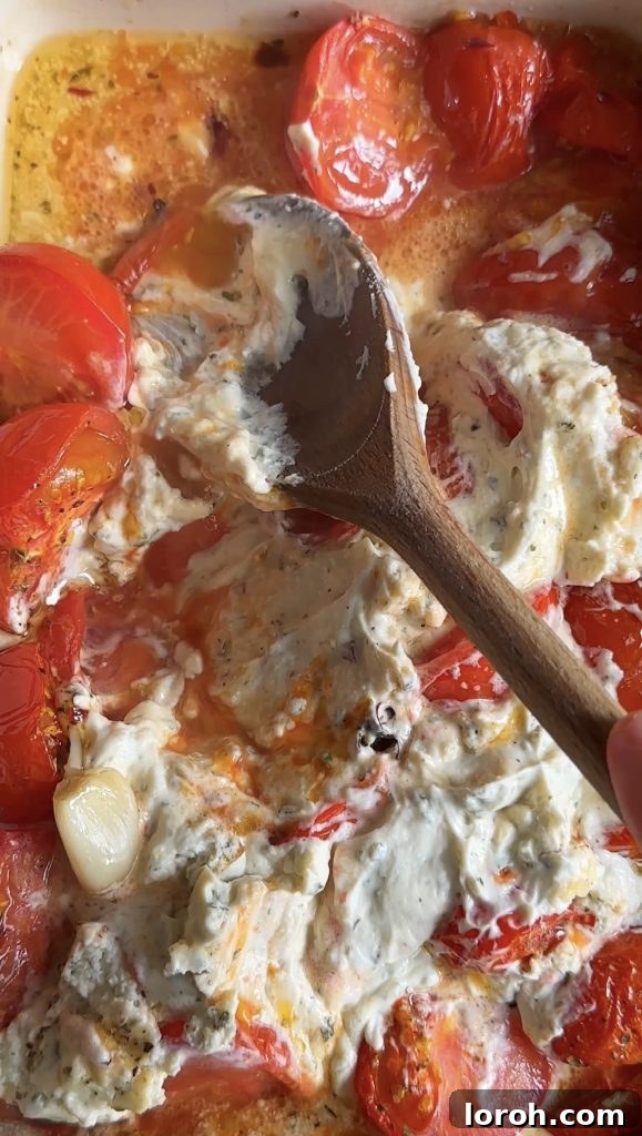 A wooden spoon mashing together roasted tomatoes, Boursin cheese, and garlic to create a creamy sauce directly in the baking dish.
