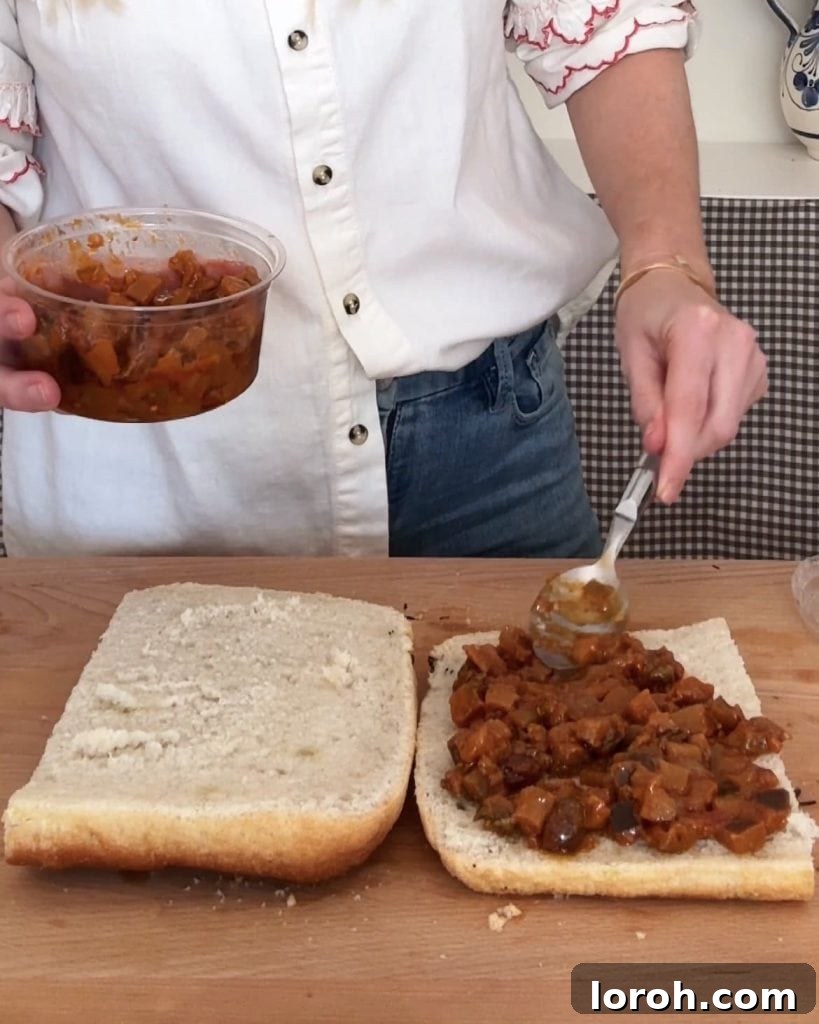 A woman spreading a generous layer of aubergine caponata onto a piece of focaccia bread with a silver spoon, preparing the sandwich base.