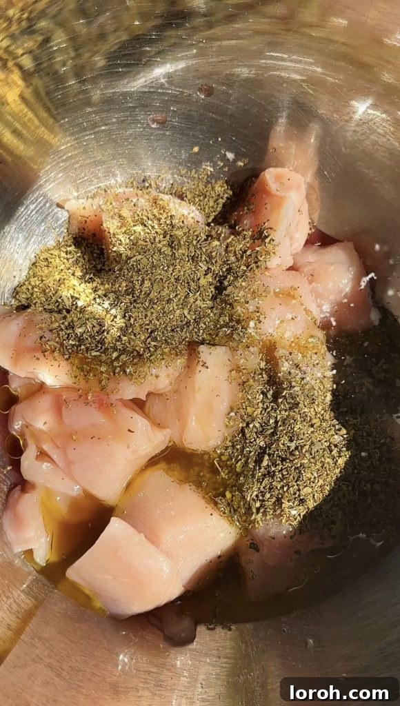 Adding dried herbs and marinade to cubed chicken breast in a metal bowl