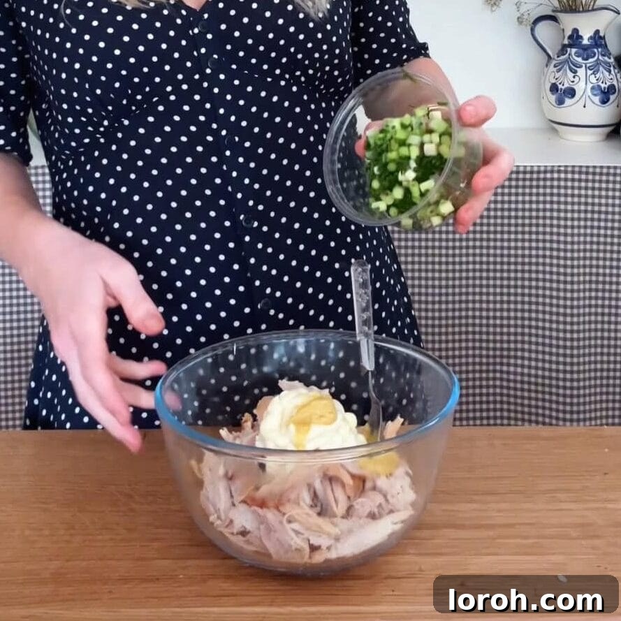 Shredded cooked chicken being mixed with mayonnaise and other ingredients in a bowl.