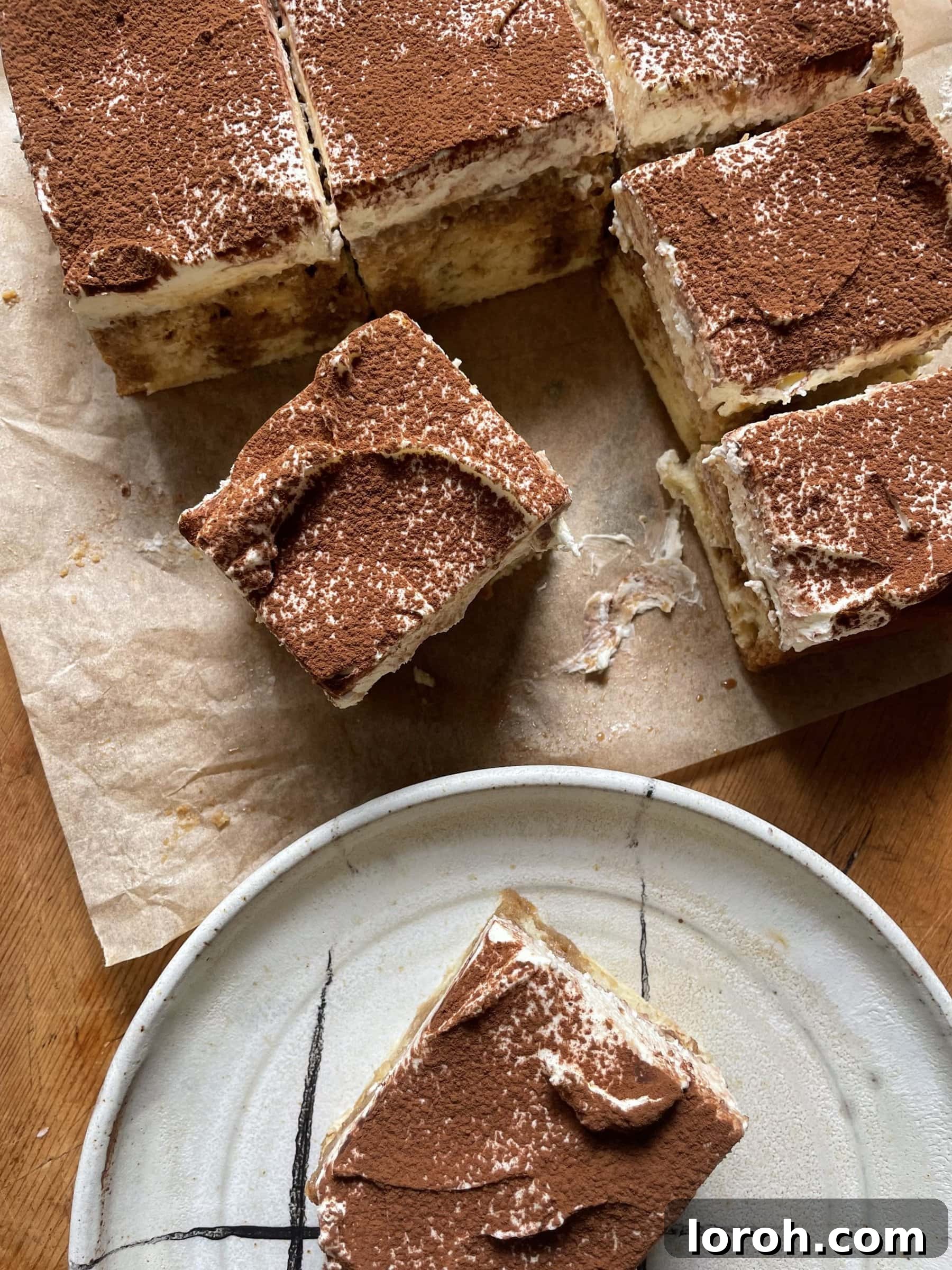 A perfectly sliced piece of Tiramisu Tres Leches Cake on a white plate, with the full cake visible in the background on a rustic wooden table, beautifully dusted with cocoa powder.