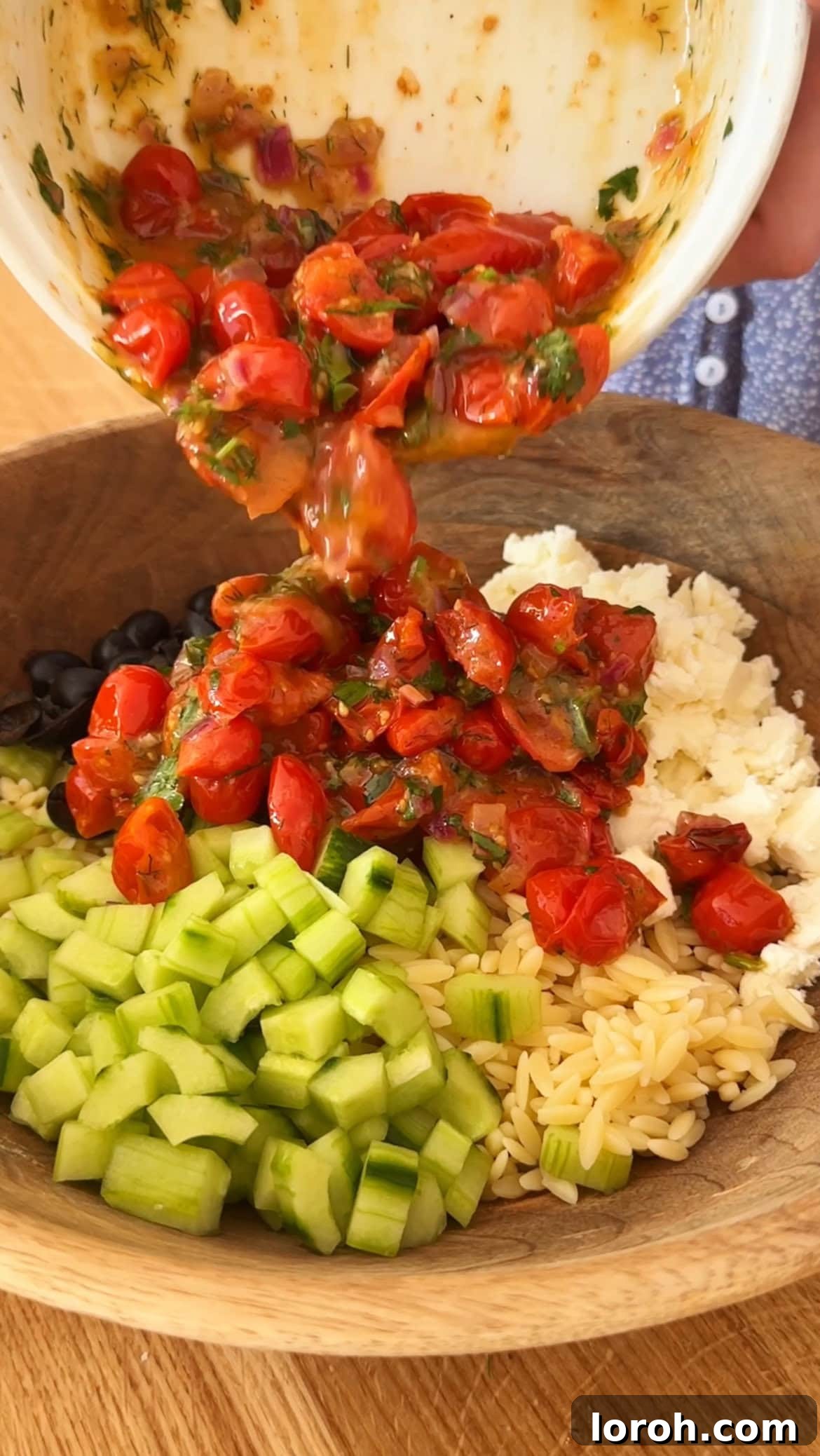 Vibrant Greek Orzo Salad 7 The completed roasted tomato and herb dressing being poured over cooked orzo, diced cucumber, olives, and crumbled feta in a large mixing bowl.