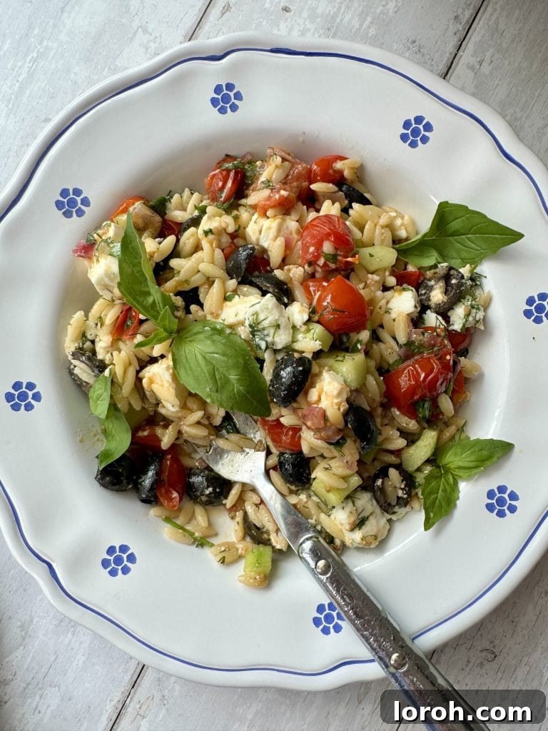 Vibrant Greek Orzo Salad 8 A close-up of Greek pasta salad in a white bowl with blue flowers, with a fork digging into the flavorful mix.