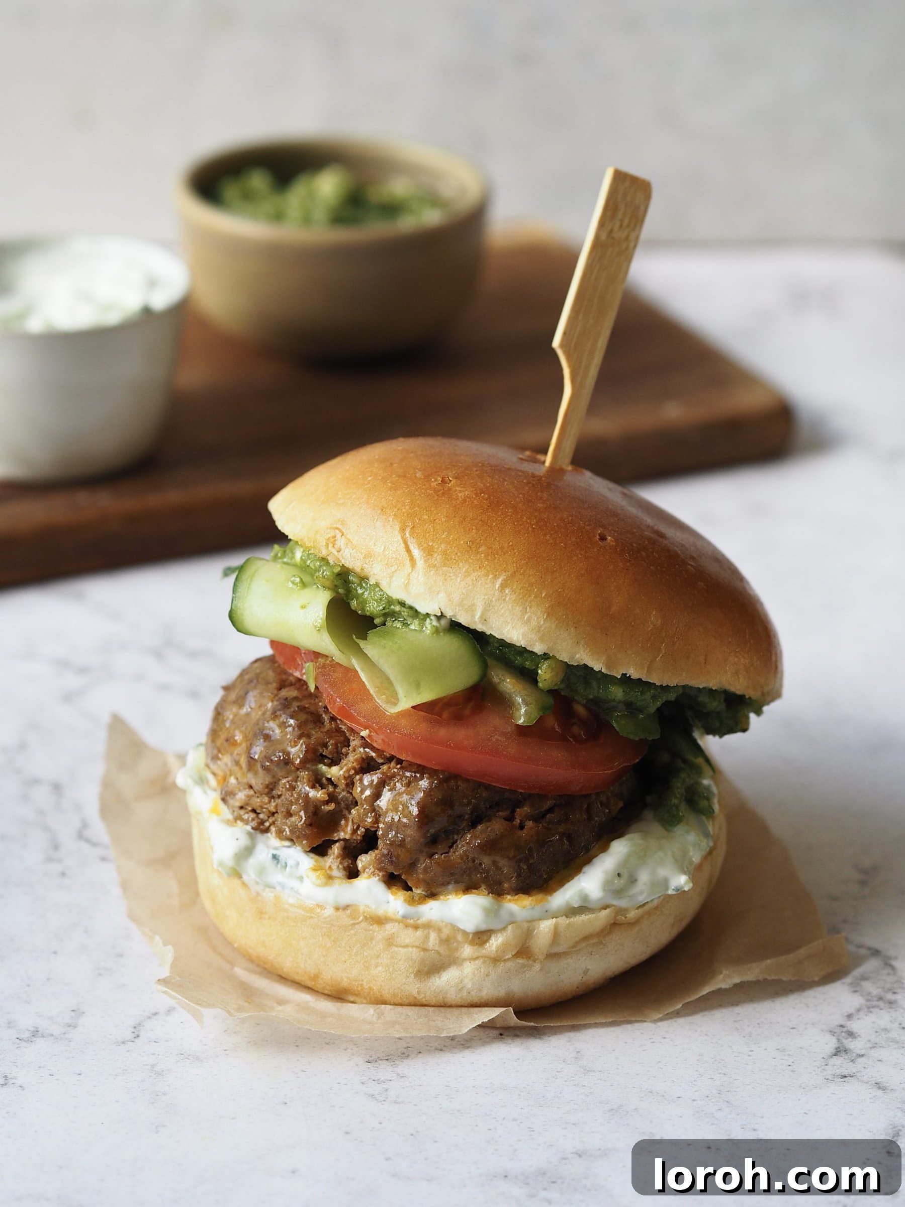 Lamb Burgers: A Fresh Mediterranean-Italian Blend 2 Lamb burger with tomato, cucumber ribbons, tzatziki and pesto on a white marble countertop.