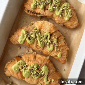 Three golden-baked pistachio croissants nestled in a baking dish, glistening with melted chocolate.