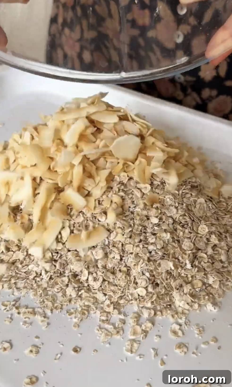 Adding coconut flakes to oats on a baking tray.