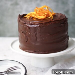 A whole Jaffa Cake Cake on a white cake stand, showcasing its dark chocolate ganache and orange zest curls.