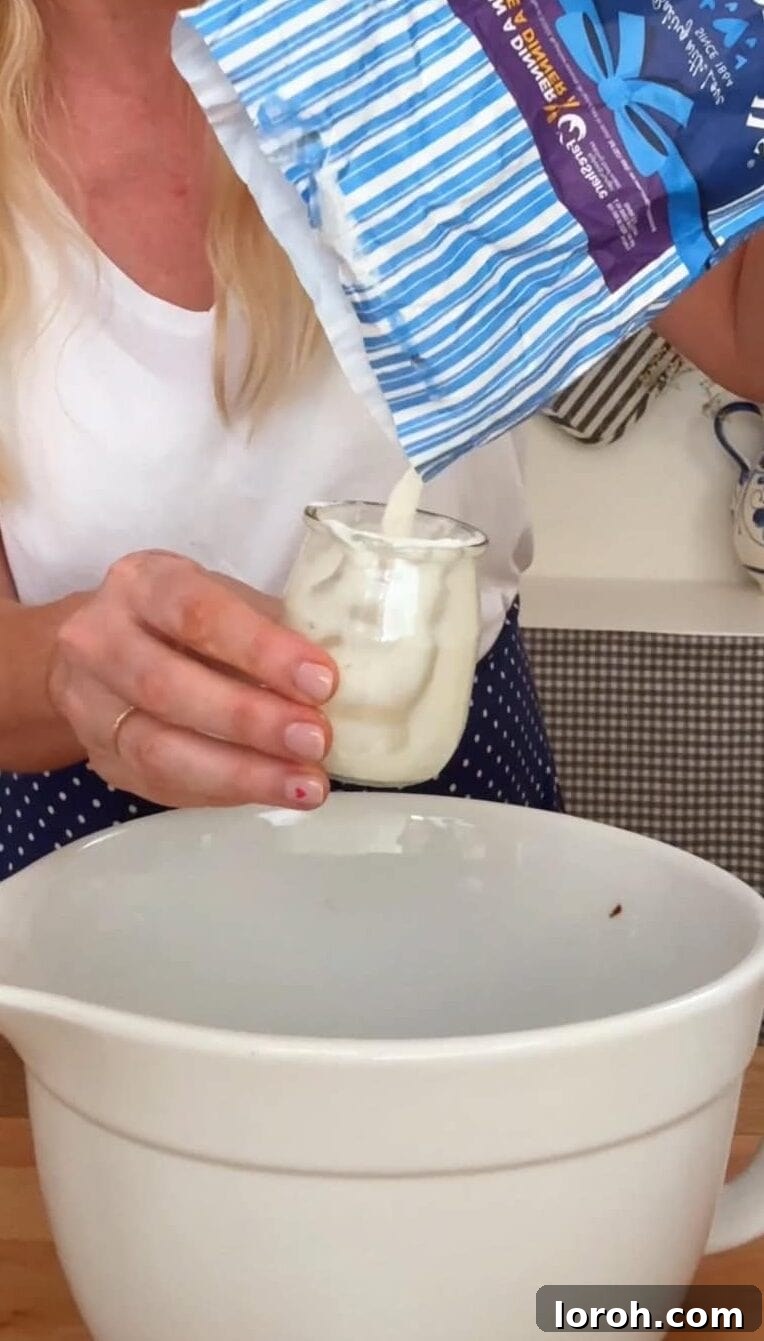French Yogurt Cake The Easiest No Measure Treat 4 A hand holding a yogurt pot, measuring out flour into a mixing bowl filled with other cake ingredients.
