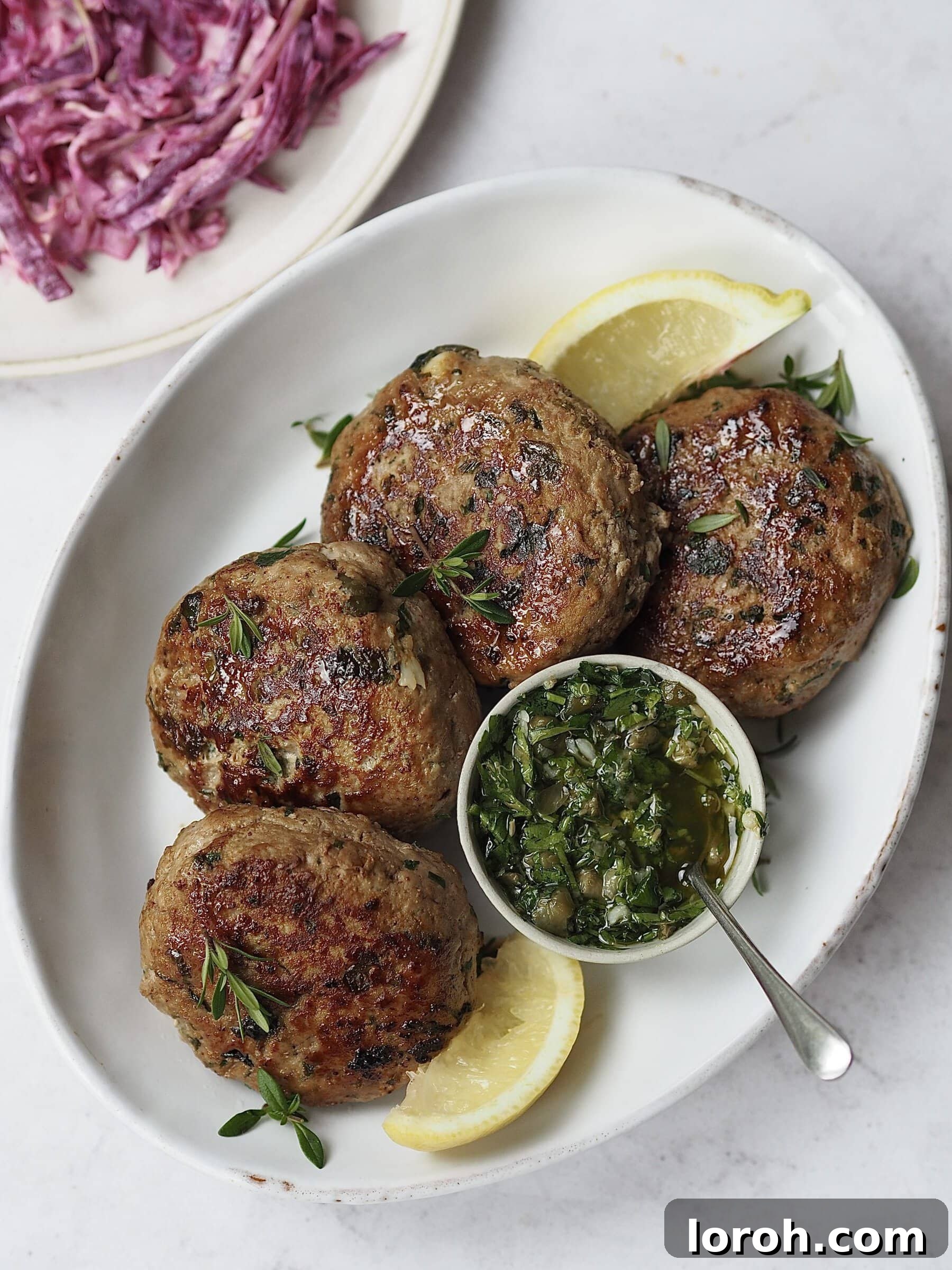 White plate of herby chicken burgers with lemon wedges, a small bowl of salsa verde and a plate of yogurt slaw on the side. A complete, healthy meal.