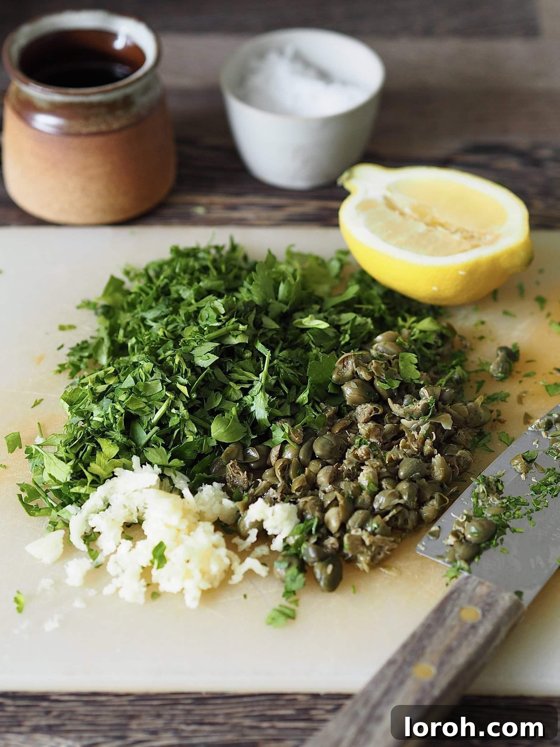 Finely chopped parsley, capers, and garlic on a wooden cutting board, ready for salsa verde preparation.