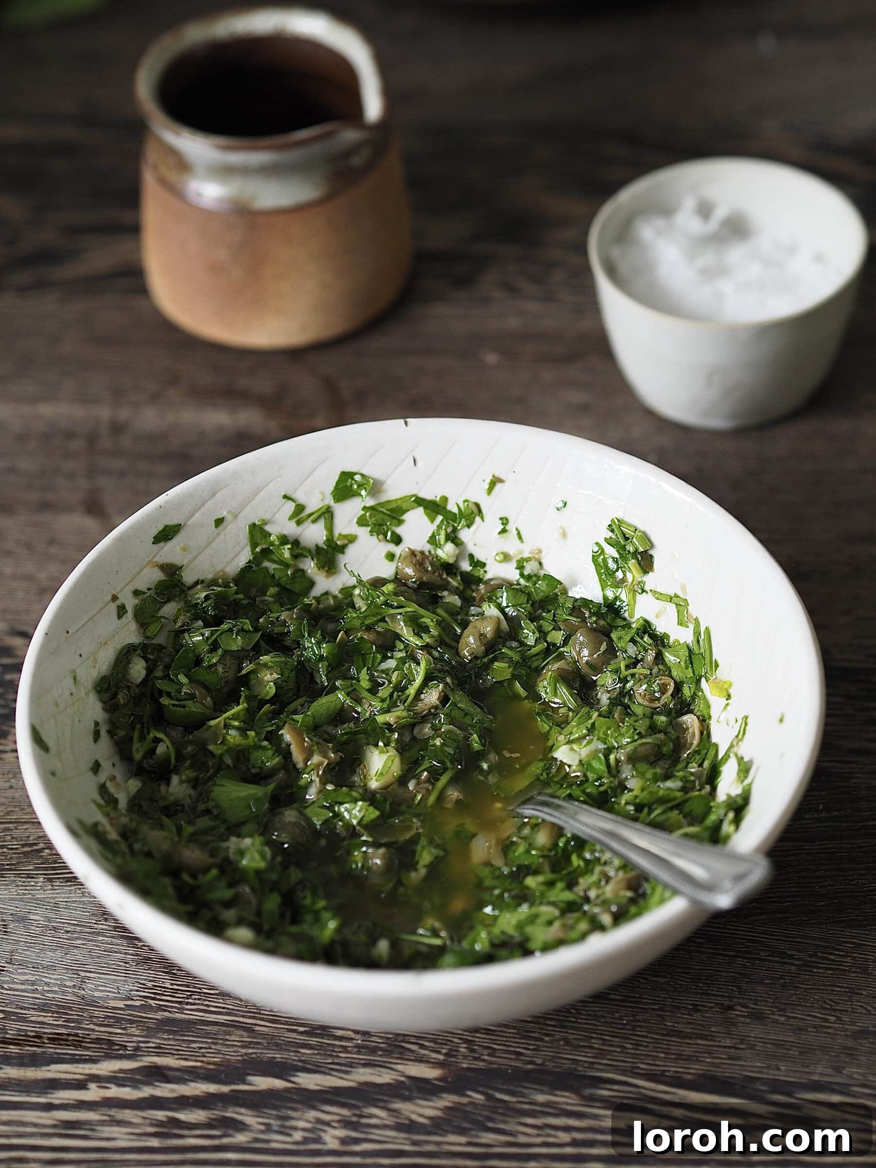 Small white bowl of vibrant green salsa verde with a teaspoon on a wooden countertop, showcasing its fresh texture.