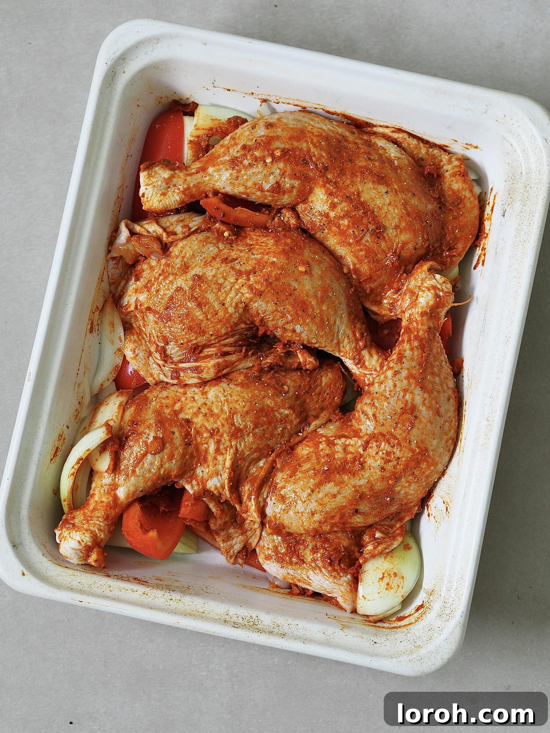 Zesty Peri Peri Chicken with Golden Potato Wedges 6 Peri peri marinated raw chicken legs in a white roasting tray on a bed of red peppers and onions.